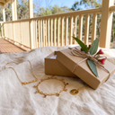 Personalised gold name necklace, birthstone charm bracelet and initial pendant on linen beside a kraft gift box and eucalyptus leaves