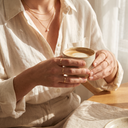 Woman in a linen shirt holding a coffee, wearing layered gold necklaces and stacked gold rings – Marliva