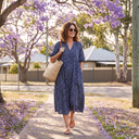 Woman in a navy floral midi dress with sunglasses and a straw tote under jacaranda trees